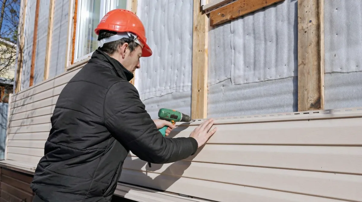 Siding replacement professional using a power drill to install new tan vinyl siding on a home exterior in Charleston, SC